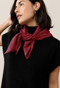 Red silk scarf tied in a bow at the neck, featuring a subtle pattern, worn over a black knitted short-sleeve top.