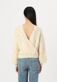 Maglione lavorato a maglia color crema con un motivo a diamante testurizzato, scollatura a V sul retro e maniche a sbuffo, abbinato a jeans a vita alta di un blu chiaro.