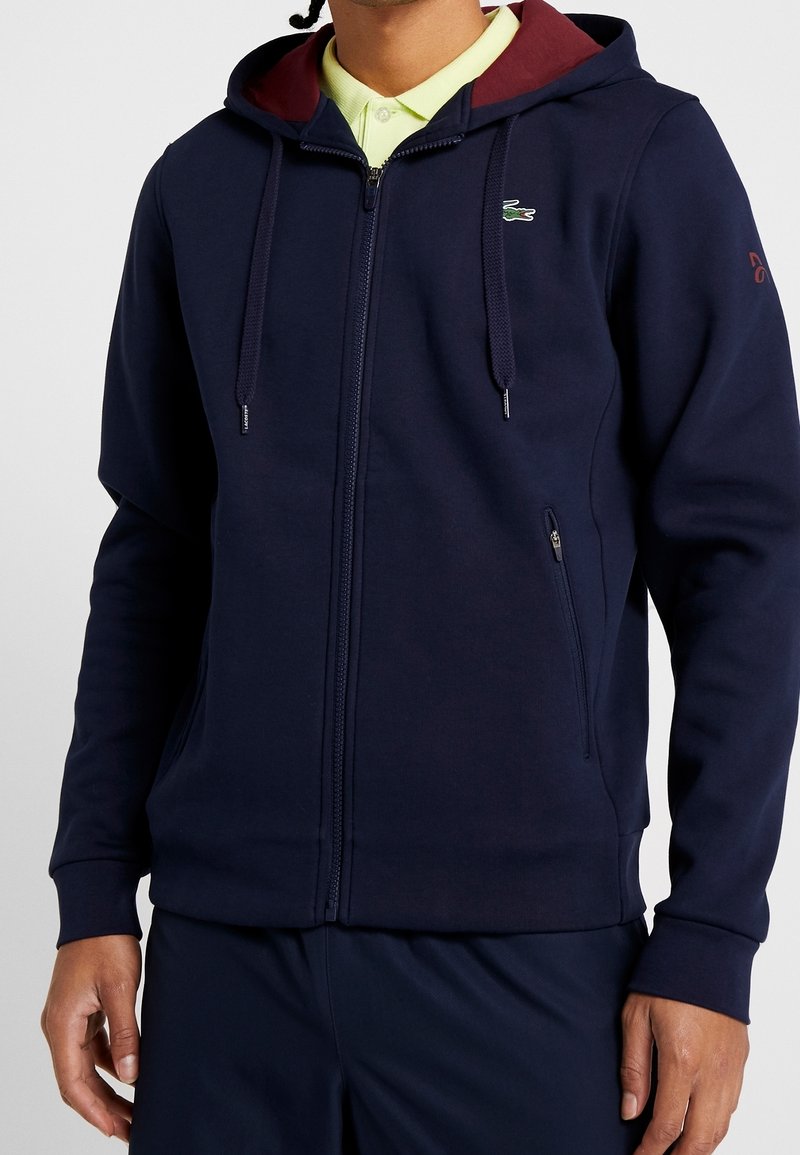 Laivastonsininen vetoketjulla varustettu huppari, jossa on viininpunainen vuorattu huppu, etuvetoketju, sivutaskut ja korostettu logo vasemmassa olkapäässä. Pehmeän tuntuinen kangas.