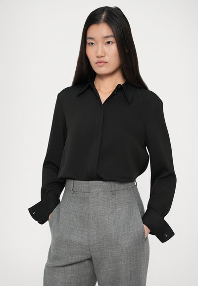 Femeie cu păr lung și drept, purtând o cămașă neagră cu nasturi bagată în pantaloni gri cu talie înaltă, având mâinile în buzunare.