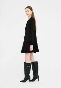 Abito mini plissettato nero con maniche lunghe e un orlo a balze a strati, abbinato a stivali in pelle nera al ginocchio con un accento in argento.