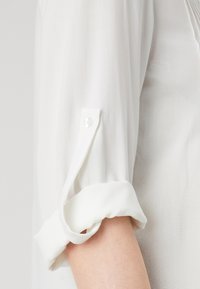 Blusa bianca a maniche lunghe con un tessuto leggermente texturizzato. Le maniche con polsini presentano un dettaglio con bottone. Design semplice e pulito, senza motivi.
