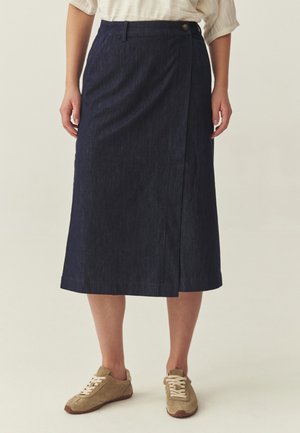 Žena v tmavomodrej sukni po kolená s prekrývajúcim sa predným dielom a béžovými teniskami stojaca pred jednoduchým pozadím.