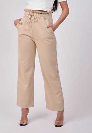 Femme portant un pantalon beige à jambes larges avec taille à cordon et poches, associé à des sandales transparentes à talons hauts et un top court blanc.