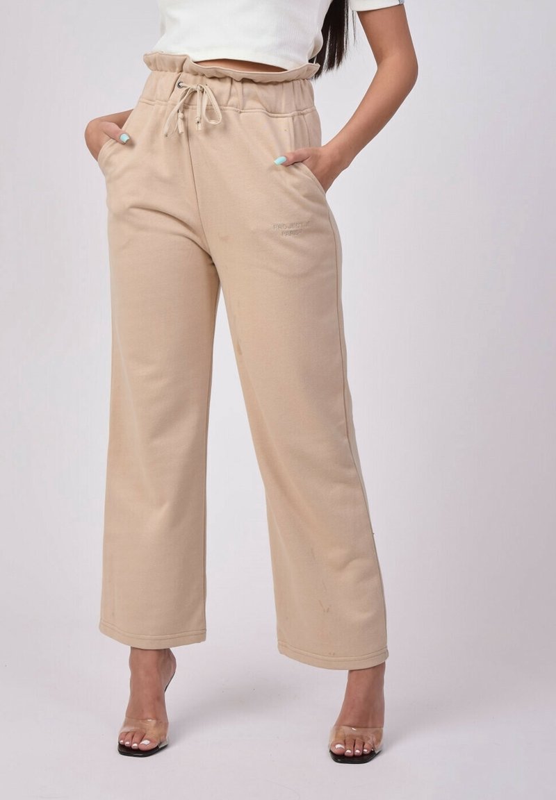 Femme portant un pantalon beige à jambes larges avec taille à cordon et poches, associé à des sandales transparentes à talons hauts et un top court blanc.