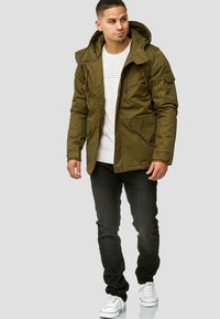 Olijfgroene parka met een capuchon, voorzien van meerdere zakken, rits- en drukknoopsluitingen, gedragen over een lichte trui, gecombineerd met donkere jeans en sneakers.