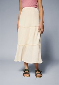 Cream-colored, textured midi skirt with horizontal seams, flared hem, and elastic waistband, paired with dark green strapped sandals.