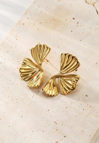 Pendientes de metal dorado con forma de hojas de ginkgo superpuestas y texturizadas sobre una superficie de piedra beige clara.