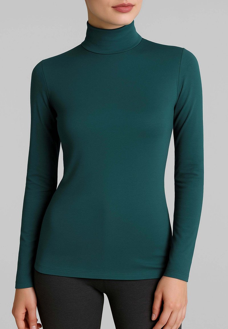 Donna che indossa un maglione attillato a maniche lunghe verde scuro con pantaloni neri su uno sfondo grigio chiaro.