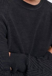 Man som bär en svart stickad tröja med texturerade mönster, armarna i kors över bröstet, synlig från halsen till midjan.