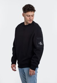 Felpa nera realizzata in tessuto morbido, con collo rotondo, maniche lunghe e una tasca sul petto con un'etichetta del logo. Indossata con jeans azzurri chiari.