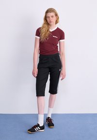 T-shirt fitted bordeaux con rifinitura bianca al colletto, pantaloni neri a vita alta con strisce bianche laterali, scarpe da ginnastica nere e calze bianche alla caviglia.