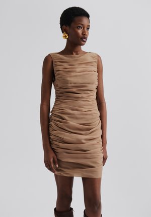 Beige, hihaton mekko, jossa on rypistyksiä ja istuva muotoilu, joka sisältää sileän pinnan ja polvipituisen helman. Kultaiset renkaat korvakorut korostavat ilmettä.