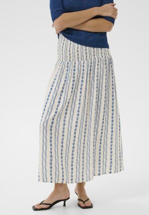 Kobieta w białej maxi spódnicy z pionowymi niebieskimi kwiatowymi paskami, niebieskiej bluzce z krótkim rękawem i czarnych sandałach na niskim obcasie, z rękami skrzyżowanymi.