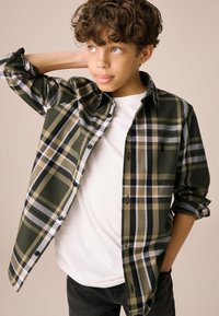 Chemise à carreaux verts à boutons avec des accents noirs et beiges, portée sur un t-shirt blanc. Un pantalon noir complète la tenue.