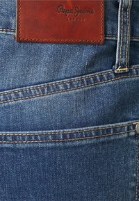Donkerblauwe denim jeans met een leren merklabel met reliëftekst, contrasterende stiksels en een detail van de voorzak.