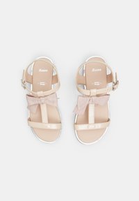 Beige sandaalit kiiltävällä pinnalla, joissa on nauhaakkents, nilkkaketju ja valkoiset pohjat. Tyylikäs muotoilu minimalistisilla yksityiskohdilla.