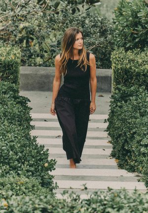 Frau in schwarzem ärmellosem Oberteil und Hose, die barfuß auf Steinstufen geht, umgeben von dichtem grünem Gebüsch im Freien.