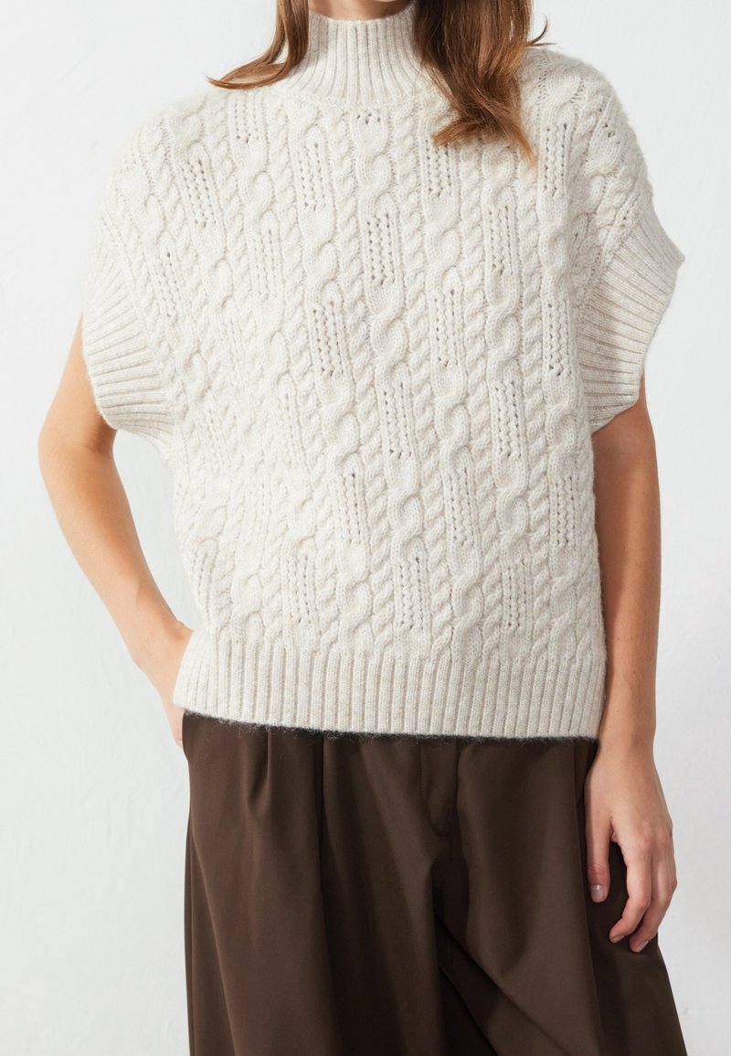 Femme portant un pull sans manches blanc en maille torsadée avec un col montant côtelé et un pantalon marron ample, mains détendues sur les côtés.