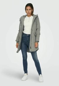 Grå hettejakke med glidelås, sidelommer, quiltet tekstur, kombinert med en hvit topp, blå jeans og hvite joggesko.