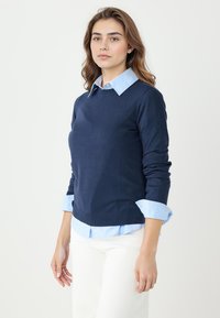 Granatowy sweter z miękkiej tkaniny z okrągłym dekoltem, noszony na jasnoniebieskiej, paskowanej koszuli. Mankiety koszuli widoczne razem z białymi spodniami.