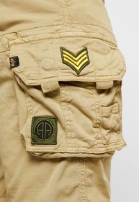 Shorts cargo beige avec des patchs verts brodés, l'un avec des chevrons et l'autre avec un logo circulaire. Le tissu est doux, avec des coutures visibles.