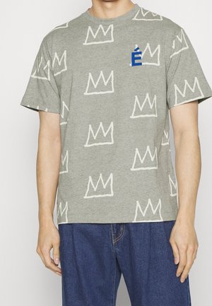 Mand iført en grå kortærmet T-shirt med hvide kronekonstruktioner og et blåt accentbogstav på brystet, kombineret med blå jeans.