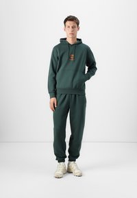 Felpa verde scuro con tasca a marsupio frontale e logo. Abbinata a pantaloni jogger coordinati, con polsini elasticizzati e vestibilità comoda.