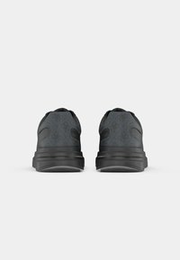 Paire de baskets gris foncé et noir vue de dos sur un fond gris clair avec des motifs subtils de logo sur le tissu.