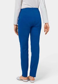 Blaue Hose mit elastischem Bund, glatter Textur, schmal zulaufendem Bein und Seitentasche. Getragen mit hellgrauen Slippern.