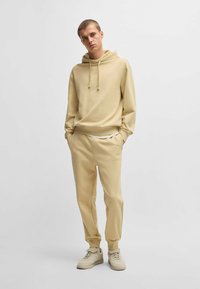 Beige hoodie met verstelbare trekkoorden, gecombineerd met bijpassende getailleerde joggers. Beide zijn gemaakt van een zachte, gestructureerde stof.
