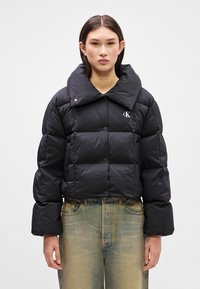 Veste rembourrée noire avec un grand col, fermeture à boutons et un logo sur la poitrine. Le matériau semble brillant ; comprend des manches larges et un design matelassé.