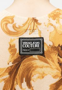 Étiquette Versace Jeans Couture à l'arrière d'un vêtement en maille beige avec un motif ornemental doré, près de l'encolure sur le haut du dos d'une personne.