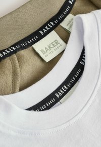 Valge puuvillane T-särgi krae, mis katab beeži kangast, millel on kontrastsed mustad sildid, kus on nähtav "BAKER BY TED BAKER" bränd.
