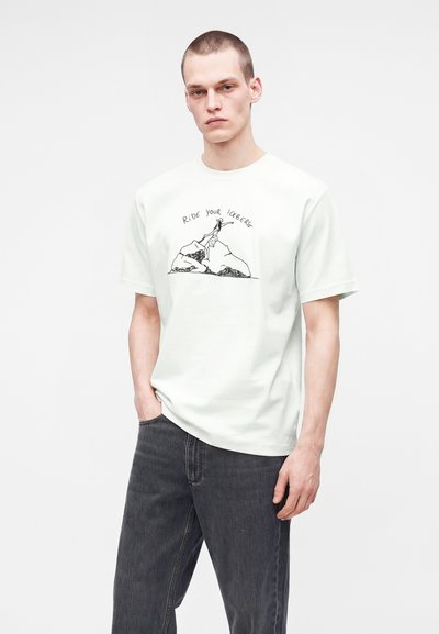 Άνδρας με κοντά μαλλιά που φοράει λευκό T-shirt με μαύρο σχέδιο γραμμής και κείμενο "Ride Your Iceberg" σε συνδυασμό με σκούρο γκρι τζιν.