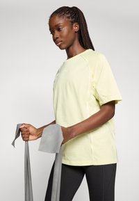 Lichtgele T-shirt met korte mouwen en witte accenten, ronde hals, gemaakt van zachte stof. Model houdt grijze weerstandsbanden vast. Zwarte leggings zichtbaar.