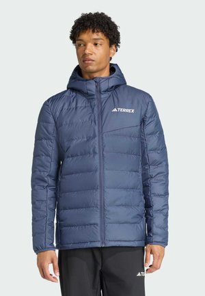 Hombre con chaqueta acolchada azul con capucha y logo TERREX, de pie con los brazos relajados, mirando ligeramente hacia la izquierda sobre un fondo claro y liso.