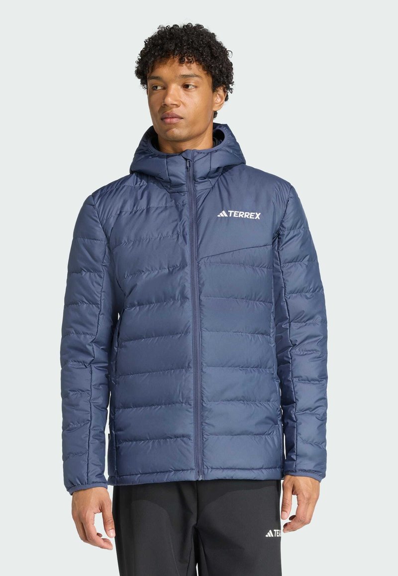 Hombre con chaqueta acolchada azul con capucha y logo TERREX, de pie con los brazos relajados, mirando ligeramente hacia la izquierda sobre un fondo claro y liso.