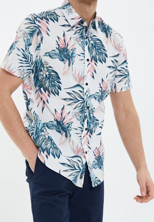 Homme portant une chemise blanche à manches courtes avec un motif de feuilles tropicales bleu et rose, associée à un pantalon bleu foncé, debout avec une main dans la poche.