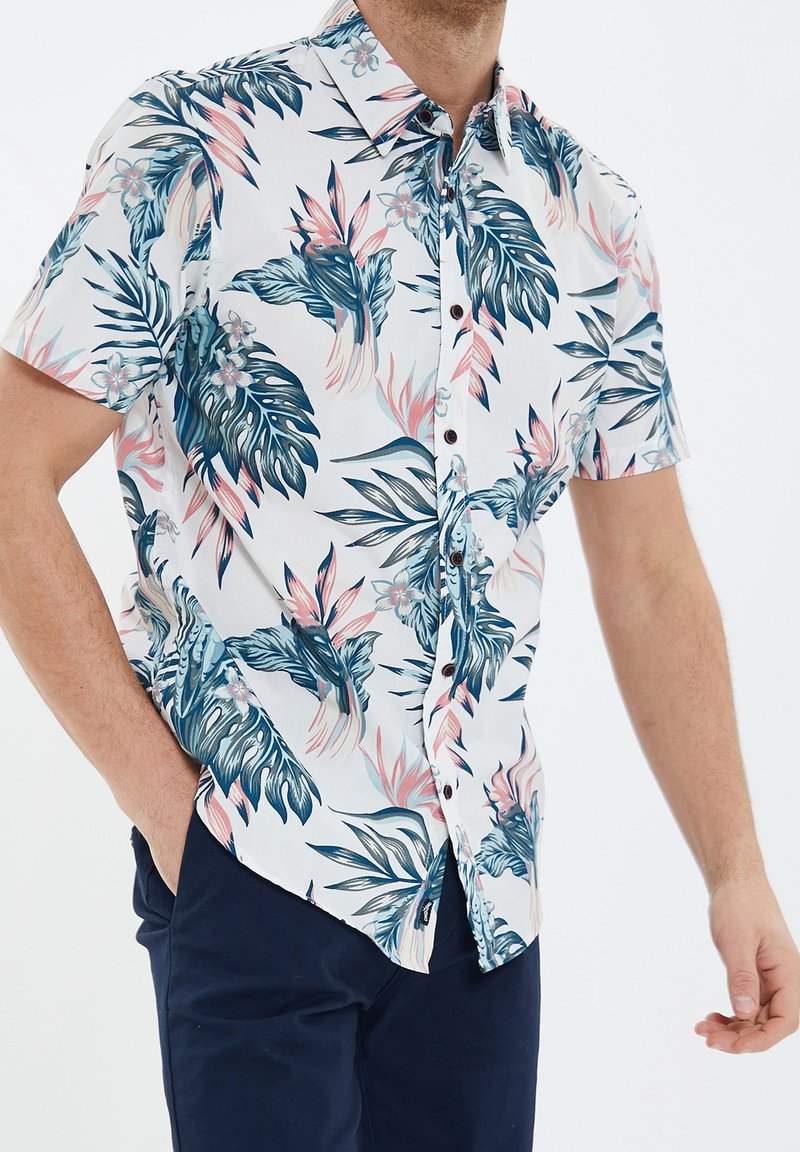 Homme portant une chemise blanche à manches courtes avec un motif de feuilles tropicales bleu et rose, associée à un pantalon bleu foncé, debout avec une main dans la poche.