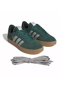 Baskets Adidas vert foncé avec semelles marron, rayures grises et blanches, et lacets gris supplémentaires à l'avant sur fond blanc.