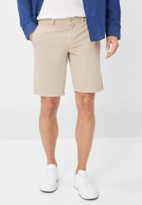 Pantaloni corti in cotone beige con vita piatta, tasche laterali e orli arrotolati, abbinati a sneakers bianche e una camicia blu.
