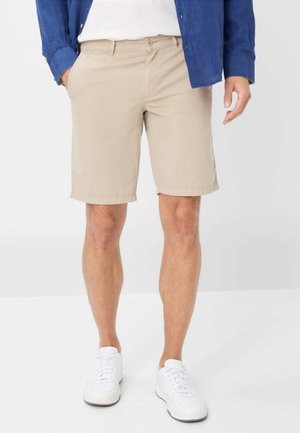 Shorts en coton beige avec un devant plat, des poches latérales et des revers, associés à des baskets blanches et une chemise bleue.