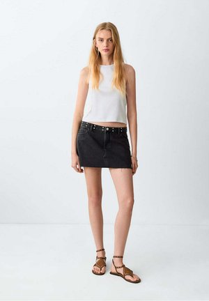 Tânără cu păr blond lung, purtând un top alb fără mâneci, fustă mini din denim negru și sandale maro cu ținte, stând în fața unui perete simplu.