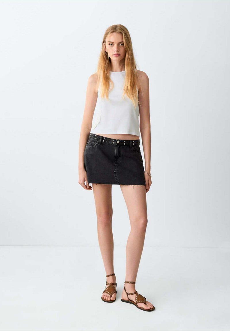 Jeune femme aux longs cheveux blonds portant un haut blanc sans manches, une mini-jupe en jean noire et des sandales cloutées marron, debout contre un mur uni.