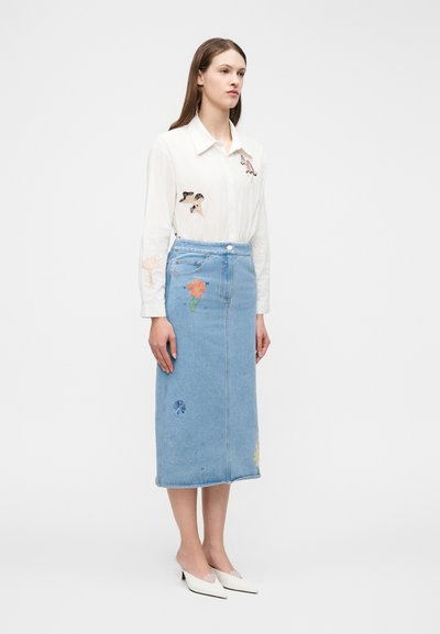 Ung kvinne iført hvit skjorte med broderi av fugl og skjell, blå denim midi-skjørt med blomsterlapper, og hvite spisse hæler, står stille.
