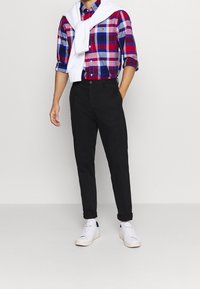 Mand iført en rød, blå og hvid ternet skjorte med oprullede ærmer, sorte bukser, hvide sneakers og en hvid sweater hængende over skuldrene.