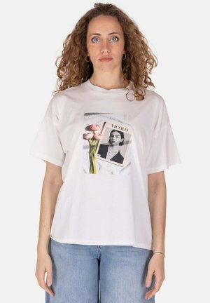 Donna con capelli ricci che indossa una t-shirt bianca con una foto di una donna e fiori rosa, abbinata a jeans azzurri chiari.