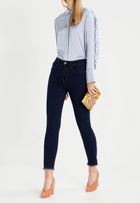 Chemise bleu clair avec des accents volantés, jeans skinny noirs, escarpins orange et pochette à motifs. Tissu lisse et coupe ajustée.