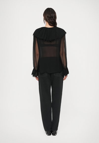 Čierna priehľadná blúzka s volánovým golierom a dlhými, voľnými rukávmi; doplnená o vysoký pás tmavých džínsových nohavíc a čierne topánky.